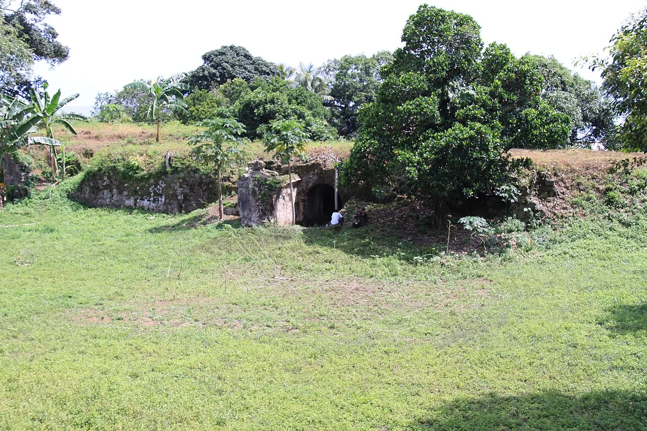 Low weathered stone walls and a dark arched entrance stand beside a grass clearing, with trees growing behind them.