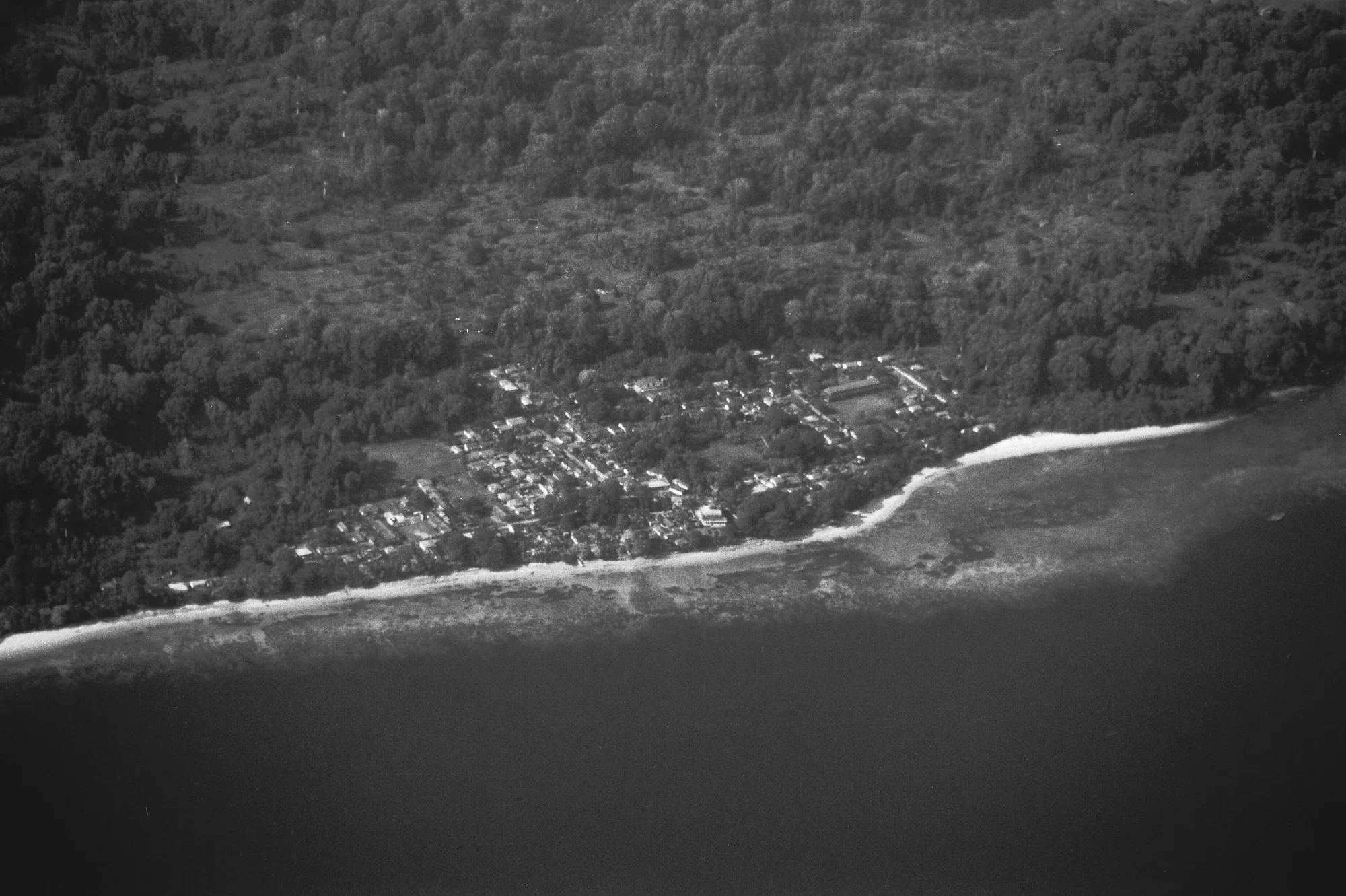 A monochrome aerial photograph shows a tree-covered coastline, clustered roofs, a pale beach and dark water.