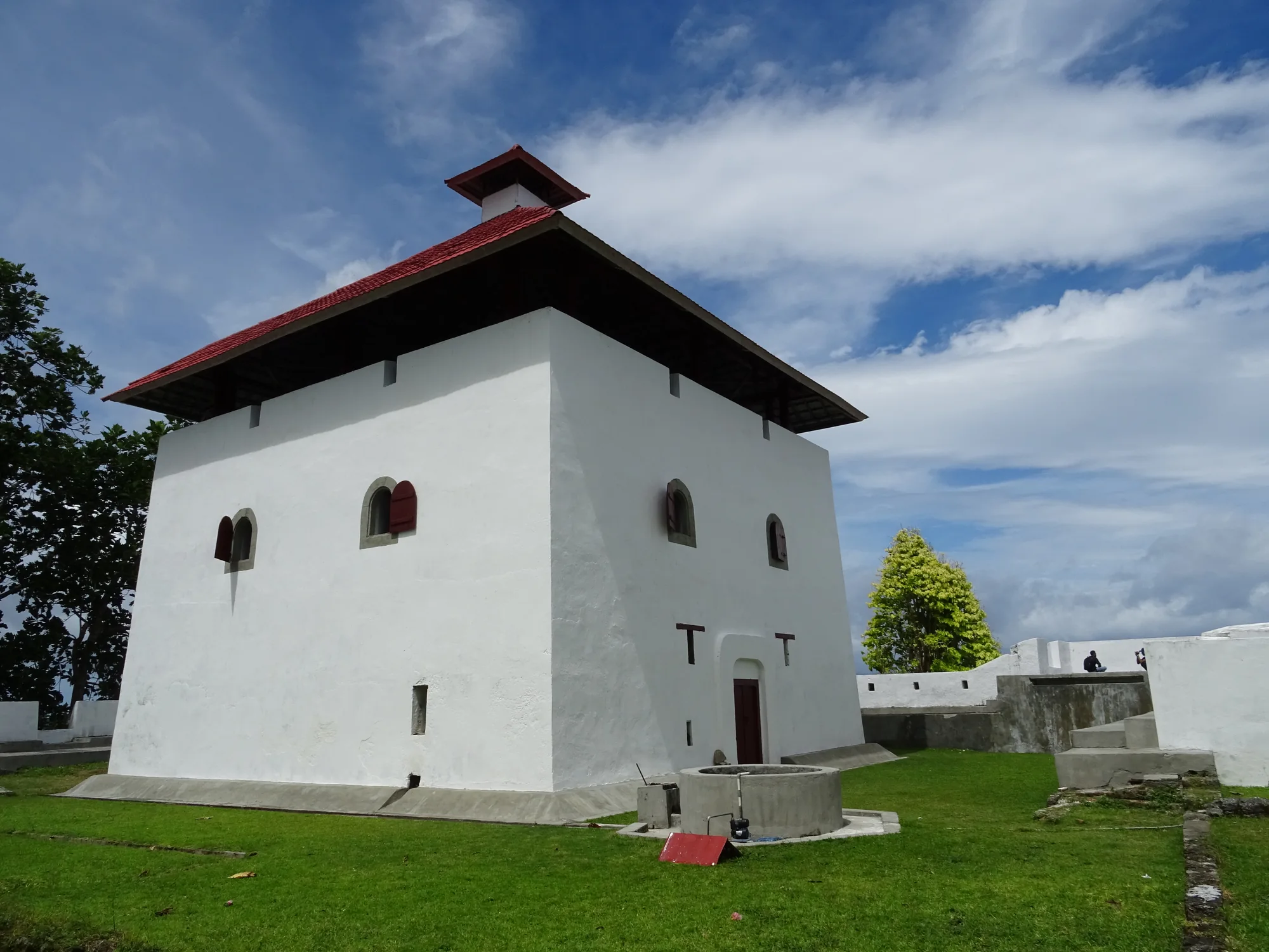 A pale masonry building with a steep red roof and low stone walls stands beside a grassy path and trees.