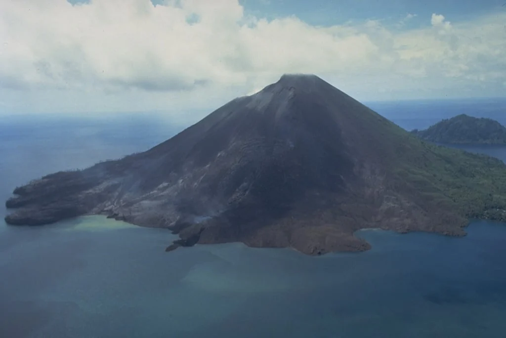 Aerial view of a largely bare volcanic cone, with dark rock extending down to blue water.