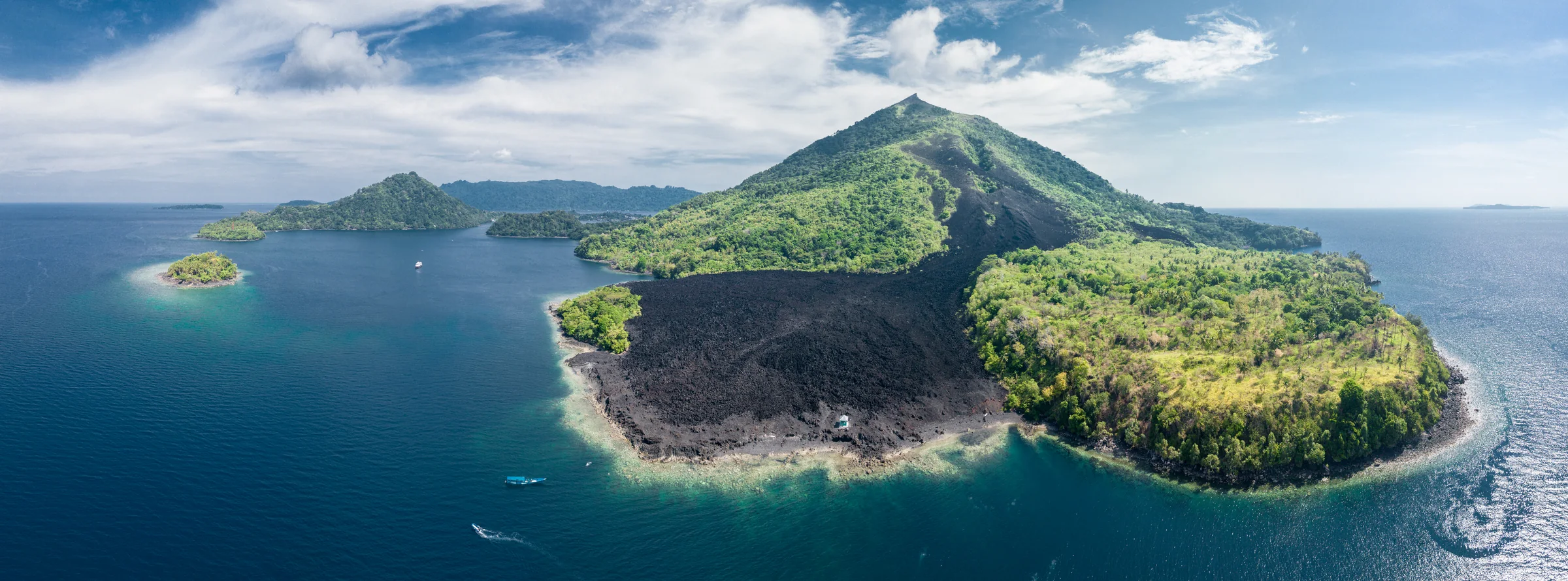 Aerial panorama of a cone-shaped island with green slopes, a broad dark strip of bare ground reaching the shoreline, and small boats offshore.