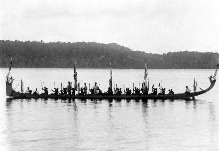 A black-and-white photograph shows a long boat with many seated people, paddles, several flags, and high curved ends against a wooded coast.