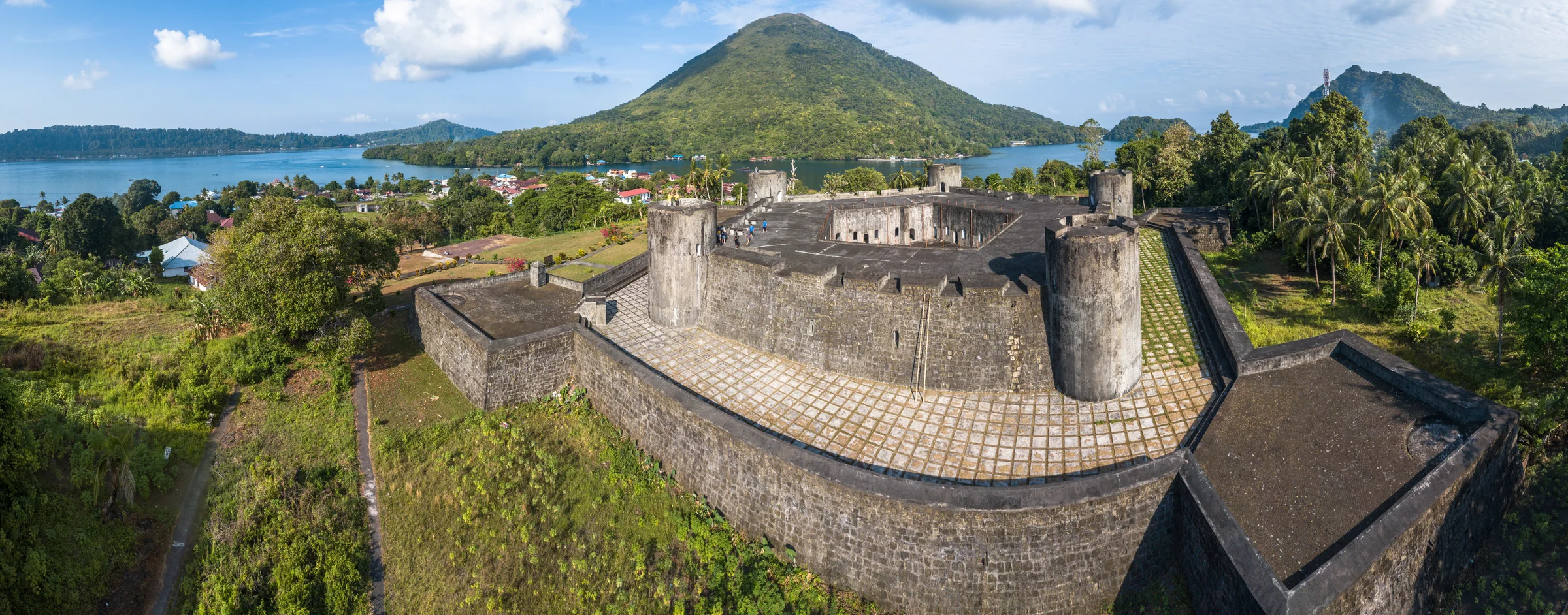A wide view of a stone fort with round corner towers, roofs and palms below it, and a cone-shaped hill across the bay.