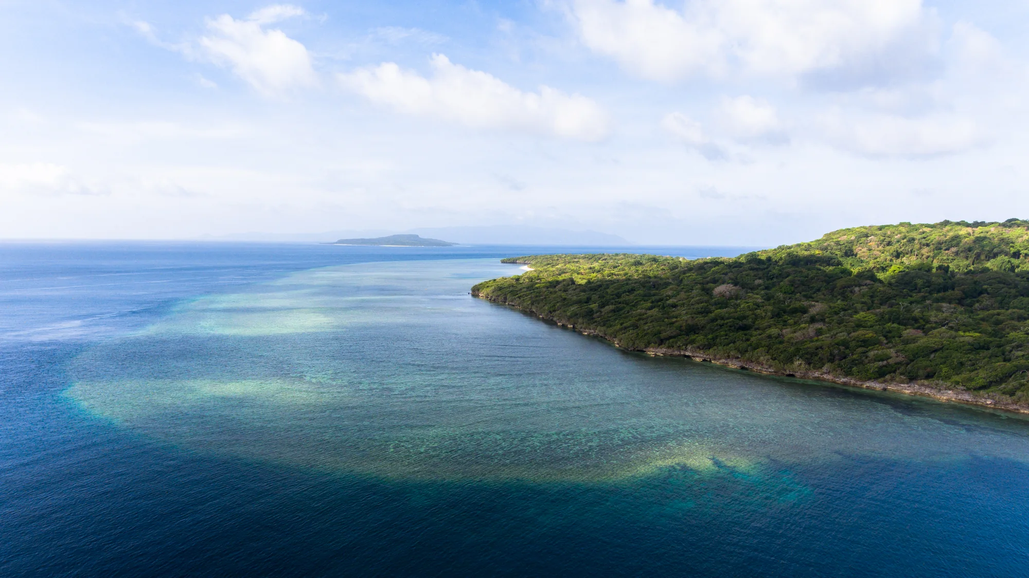 A forested coastline curves beside bands of pale turquoise water and deeper blue sea, with low islands on the horizon.